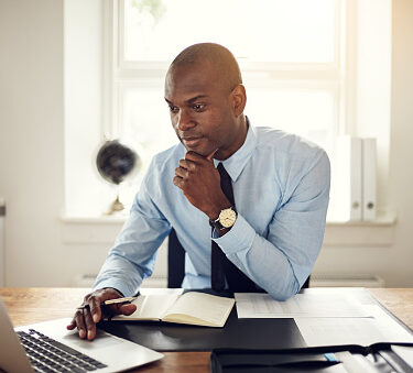 Young African business executive wearing a shirt and tie sitting at his desk in an office working online with a laptop