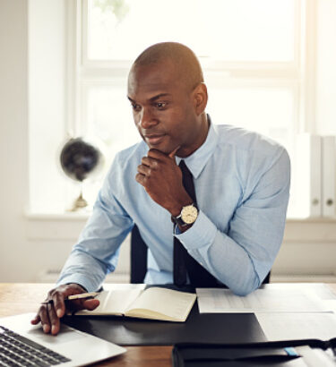 Young African business executive wearing a shirt and tie sitting at his desk in an office working online with a laptop
